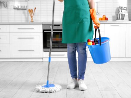 close shot of a woman standing with a bucket and a mop in her hand , also wearing cleaning gear.